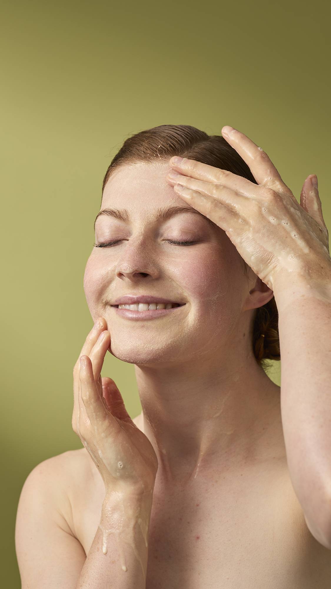 A person with their eyes closed and a gentle smile massages a foamy cleanser into their face. Their wet hands are covered in light soap suds, and they stand against a solid olive-green background.