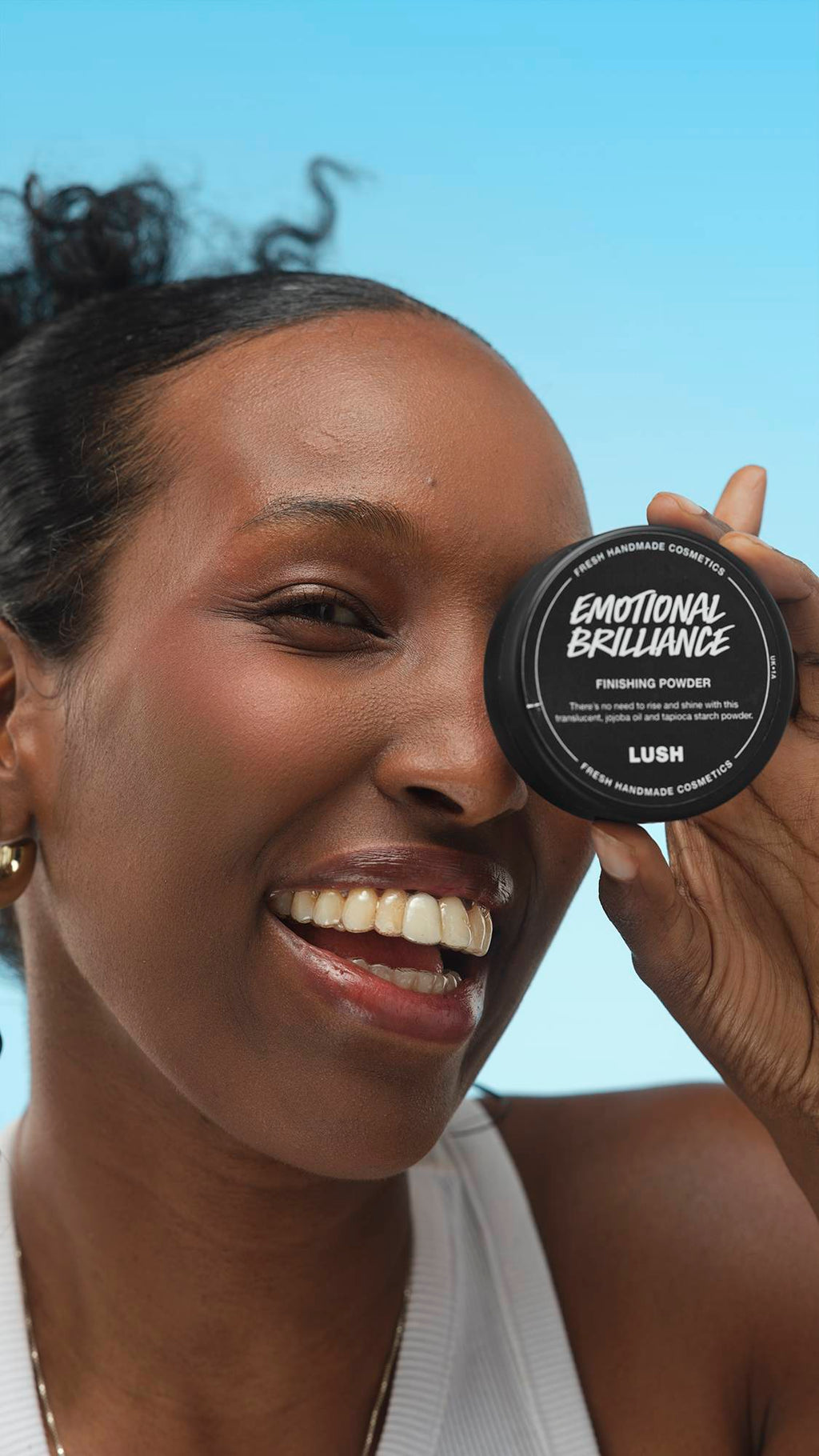 A person with dark skin smiles widely, holding the black pot of Emotional Brilliance ~ Finishing Powder over their left eye. They wear a white top and a gold hoop earring, against a bright blue background. The text on the pot is clearly visible.