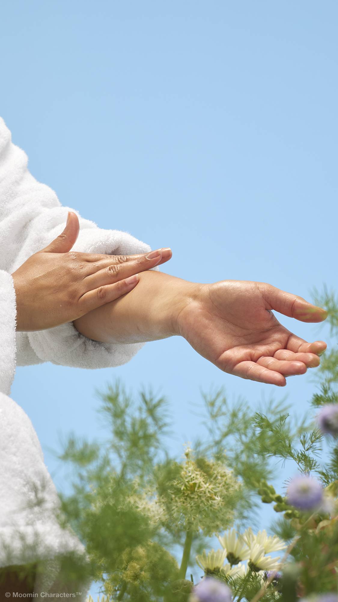 A close-up shot of a person in a white dressing gown smoothing the cream-coloured "Moominmamma" lotion onto their forearms and hands. Soft green foliage and a white flower are visible in the background against a clear blue sky.