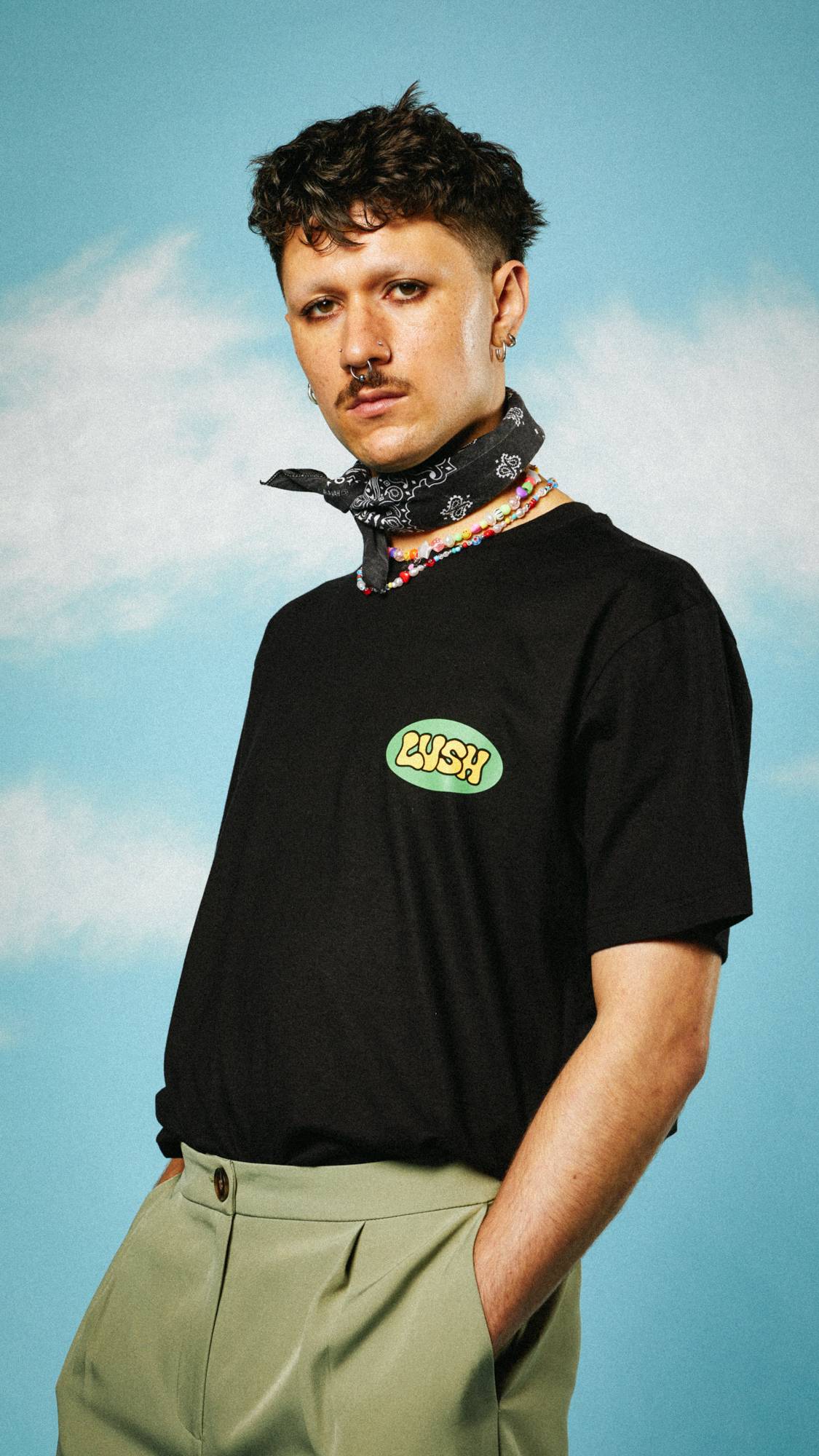 Model is stood wearing camo-green trousers and a patterned neck scarf with the LUSH Retro Bubble t-shirt.